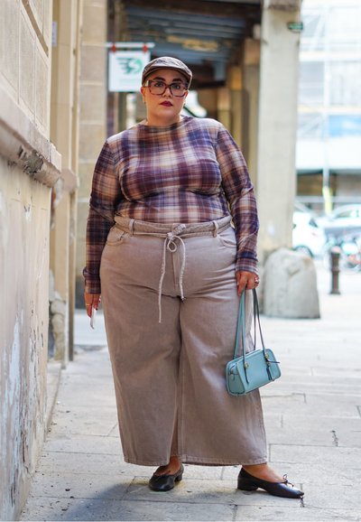 Camisa de manga larga a cuadros en púrpura y beige, combinada con pantalones anchos beige y bailerinas negras. Pequena bolsa azul, fondo claro texturizado.