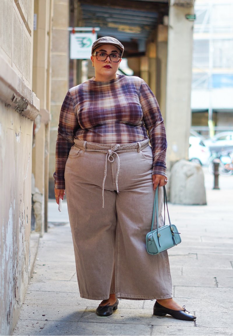 Chemise à manches longues à carreaux en violet et beige, associée à un pantalon large beige et des ballerines noires. Petit sac à main bleu, arrière-plan texturé clair.