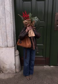 Veste en cuir marron, jean large bleu et sac à main en daim beige. Tient un bouquet de baies rouges et de feuillage vert emballé dans du papier kraft.
