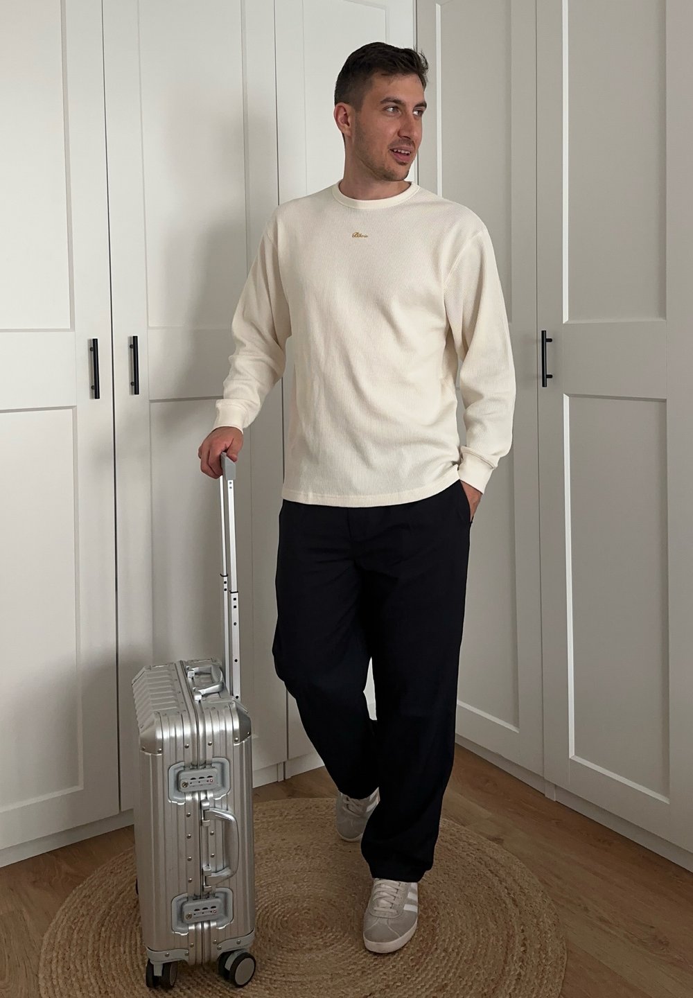 Valise rigide argentée avec des rainures, roues roulantes et poignée télescopique. Modèle portant un pull crème texturé et un pantalon foncé.