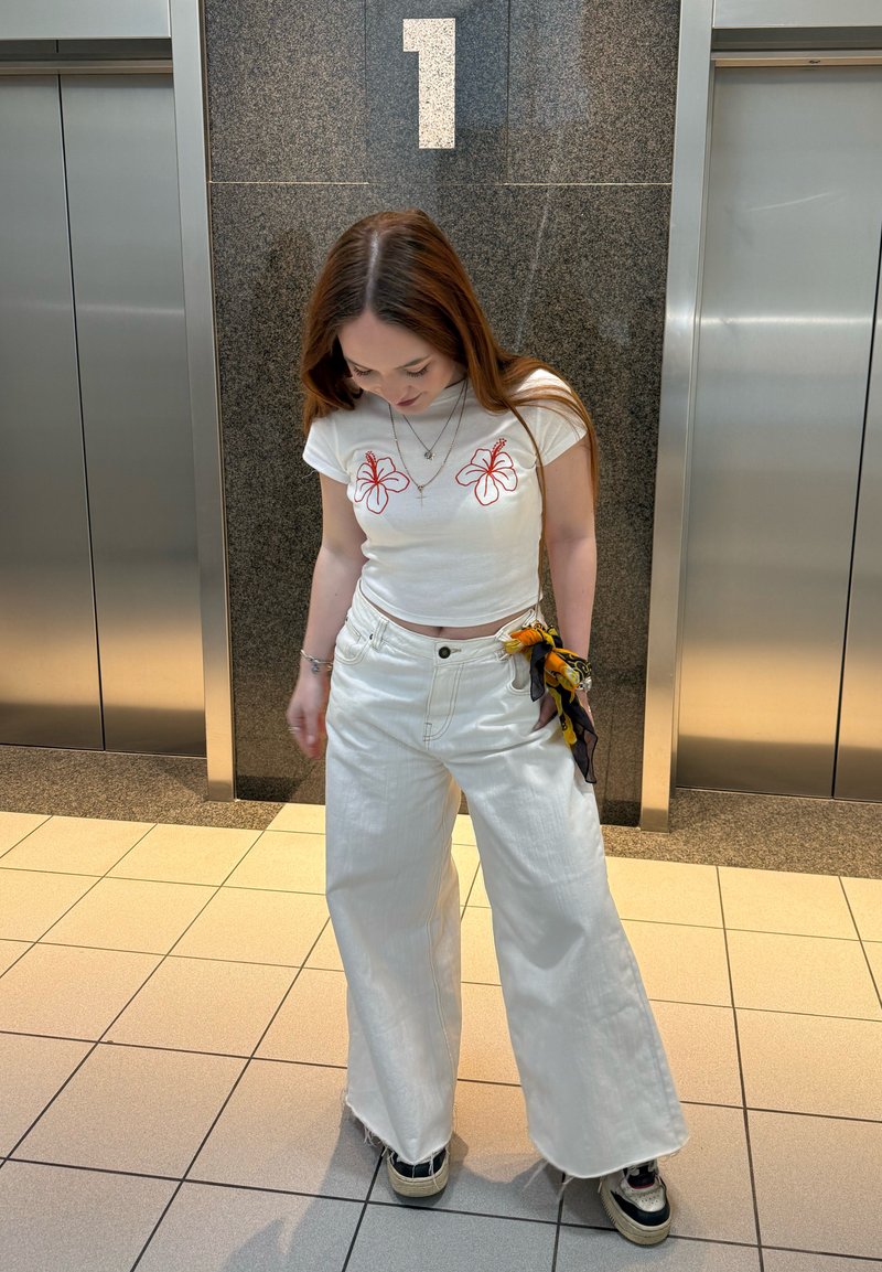 Jeune femme en pantalon blanc à jambes larges et crop top avec des fleurs rouges, debout devant les portes de l’ascenseur sous un grand panneau avec le numéro 1.