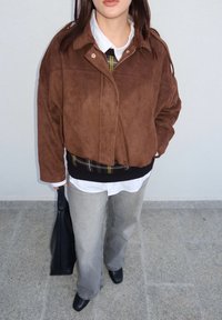 Veste en daim marron avec un col, portée par-dessus une chemise blanche et un pull noir. Un jean gris à jambes larges et des chaussures noires complètent la tenue.