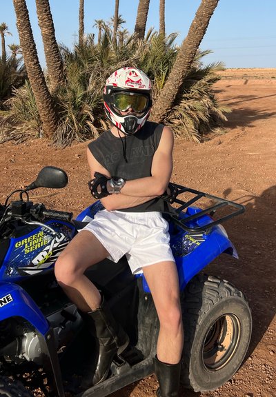 Vehículo todoterreno azul con neumáticos negros, con un marco protector. Una figura que lleva un casco blanco, visera ahumada y guantes negros se sienta cerca.