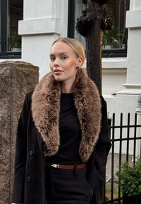 Femme aux cheveux blonds lissés en arrière, portant un manteau noir avec un grand col en fourrure marron, debout en plein air près d'un pilier en pierre et d'une clôture en métal.