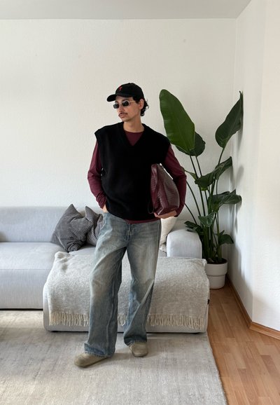 Hombre con chaleco negro, camiseta de manga larga burdeos, vaqueros, gorra y gafas de sol sostiene una bolsa grande marrón de pie en una sala de estar minimalista con plantas.