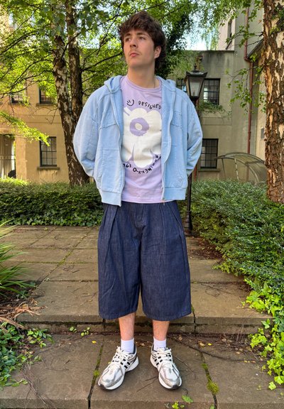 Sudadera con cremallera azul claro sobre una camiseta lila con un diseño floral, combinada con pantalones cortos de mezclilla oscuros y suéteres deportivos plateados.