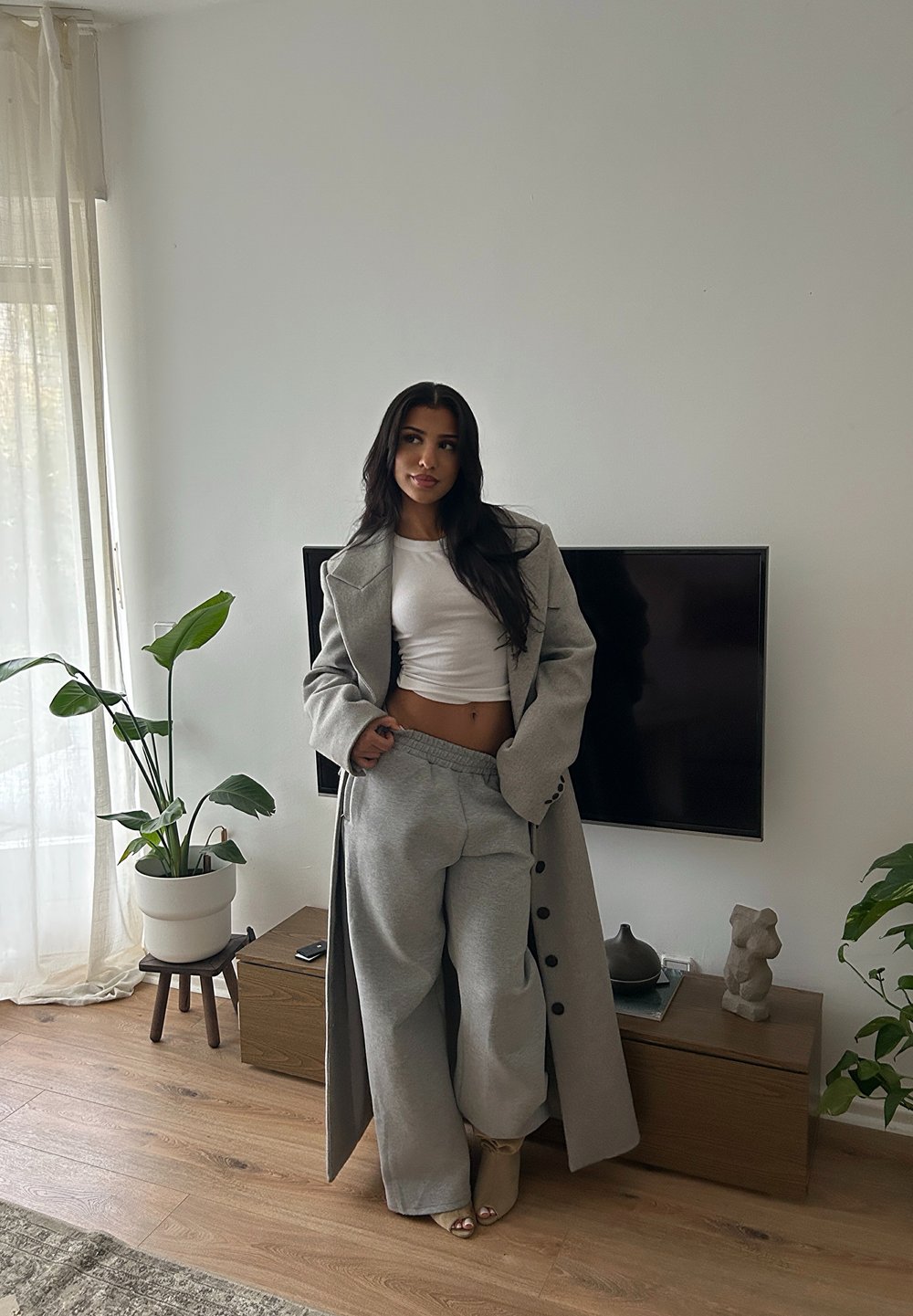 Kvinna med långt mörkt hår som bär en grå kappa, vit kort topp och grå byxor, stående i ett vardagsrum med växter och TV på väggen.