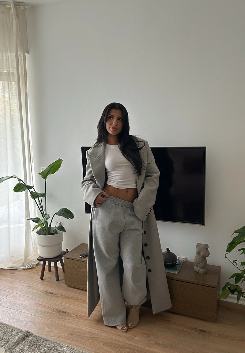 Naisel, kellel on pikad tumedad juuksed, on seljas hall mantel, valge lühike topp ja hallad püksid, seistes elutoas, kus on taime ja seinal TV.