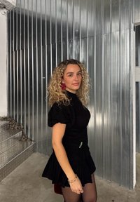 Svart kjole med puffermer, tettsittende liv og flagrende skjørt, prydet med en grønn dekorativ detalj. Krøllete hår, røde øredobber, som står mot en metallvegg.