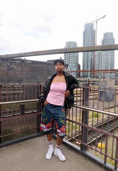 Chaqueta bomber negra, top sin mangas rosa, pantalones cortos de denim azul con patrones florales, zapatillas blancas y calcetines a rayas, de pie sobre una plataforma.