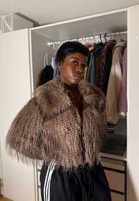 Personne portant une veste marron à plumes se tient devant une armoire ouverte avec différentes vestes et manteaux suspendus à l'intérieur.