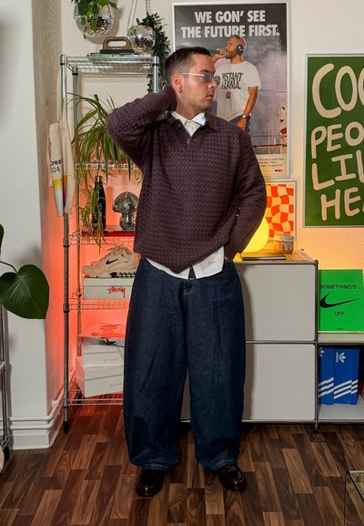 Suéter de punto morado de gran tamaño sobre una camisa blanca de cuello, combinado con pantalones vaqueros oscuros de corte suelto y zapatos negros, en un interior moderno.