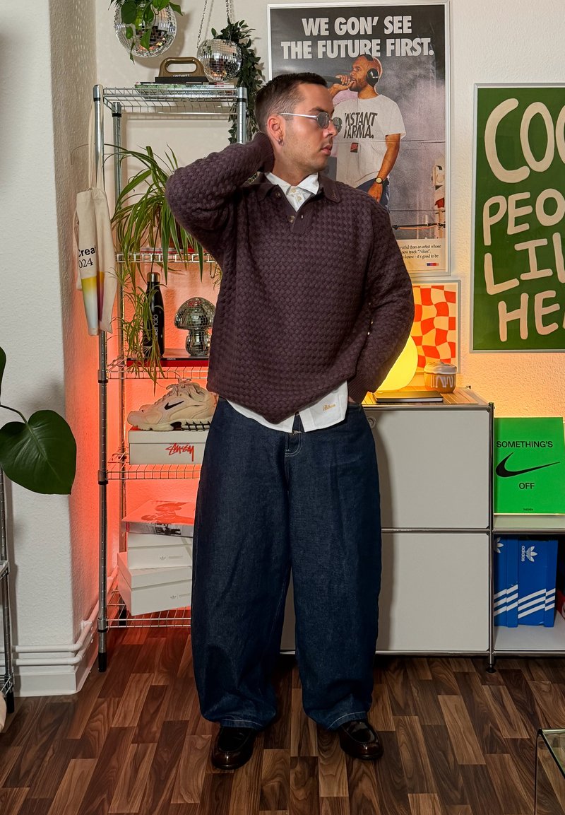 Oversized purple knit sweater over a white collared shirt, paired with loose-fitting dark denim trousers and black shoes, set in a modern interior.