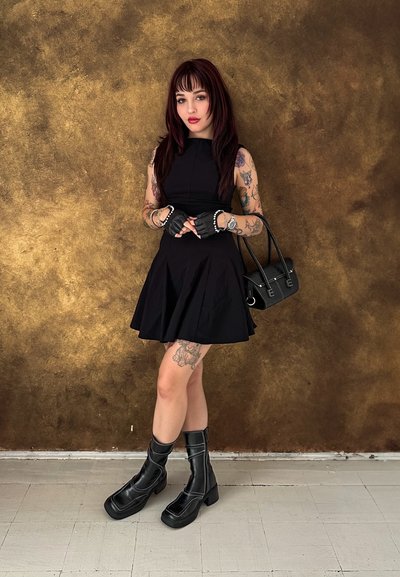 Vestido negro sin mangas con falda acampanada, combinado con botas de combate negras con cremalleras. Los accesorios incluyen guantes sin dedos y un bolso negro.