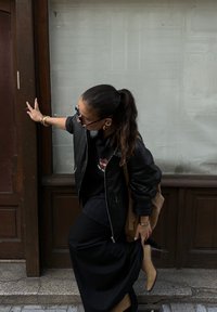 Μαύρη δερμάτινη βομβιστική ζακέτα πάνω από μαύρο μπλουζάκι, συνδυασμένη με μακριά μαύρη φούστα και καφέ μπότες μέχρι τον αστράγαλο. Ορατή μπεζ τσάντα και αξεσουάρ.