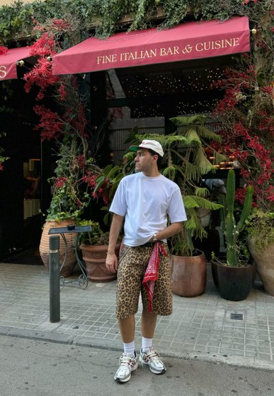 Shorts de leopardo, camiseta blanca, bandana roja y zapatillas blancas. Rodeado de plantas en macetas y un toldo rojo con texto.