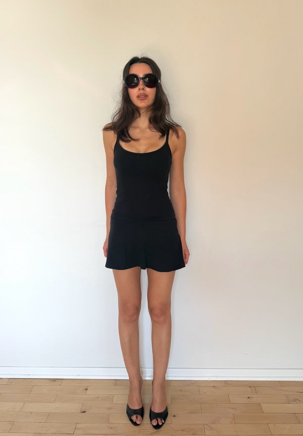 Vrouw staat tegen een beige muur, draagt een zwart tanktopje, een zwarte rok, grote donkere zonnebril en zwarte open schoenen.
