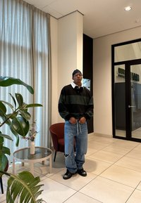 Mann iført en svart og grønn stripete genser, revne blå jeans, svarte støvler og et hodeplagg står i et moderne rom med planter og en glassdør.