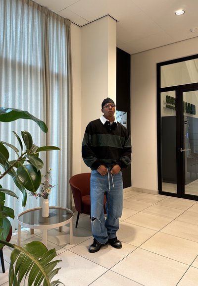 Hombre vestido con un suéter a rayas negro y verde, jeans azules rasgados, botas negras y un pañuelo en la cabeza, se encuentra en una habitación moderna con plantas y una puerta de cristal.