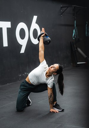 Kettlebell con acentos azules sostenido por encima en un gimnasio. Sujeto con camiseta blanca ajustada y pantalones verde oscuro, realizando una zancada sobre un suelo negro.