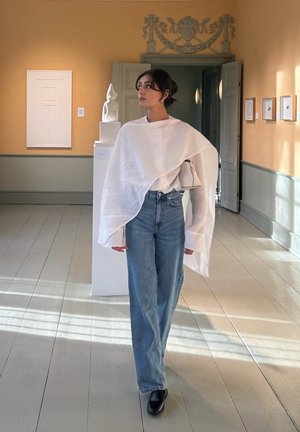 Mujer con top blanco de gran tamaño y vaqueros azules está en una galería de arte con una escultura y obras de arte enmarcadas en las paredes.