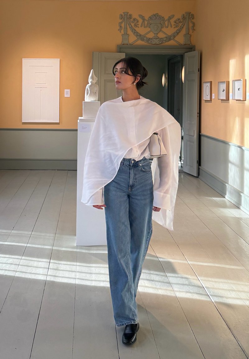 Γυναίκα που φοράει λευκό φαρδύ τοπ και μπλε τζιν στέκεται σε γκαλερί τέχνης με γλυπτό και κορνιζαρισμένα έργα τέχνης στους τοίχους.