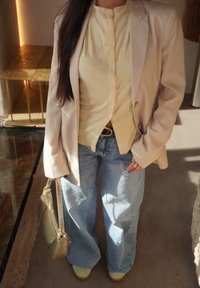 Beige blazer over en ribbet, lys gul topp, sammen med vide denimjeans og pastellgrønne sneakers. Holder en teksturert gullveske.