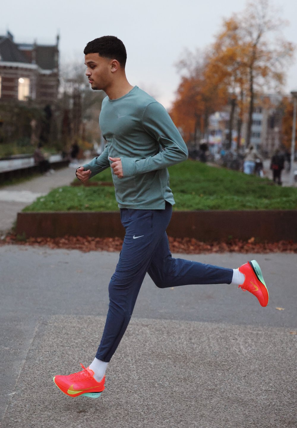 Tenue de sport comprenant un haut à manches longues vert clair et un pantalon de jogging bleu marine. Baskets orange vif avec des accents turquoise. Chemin texturé et végétation.