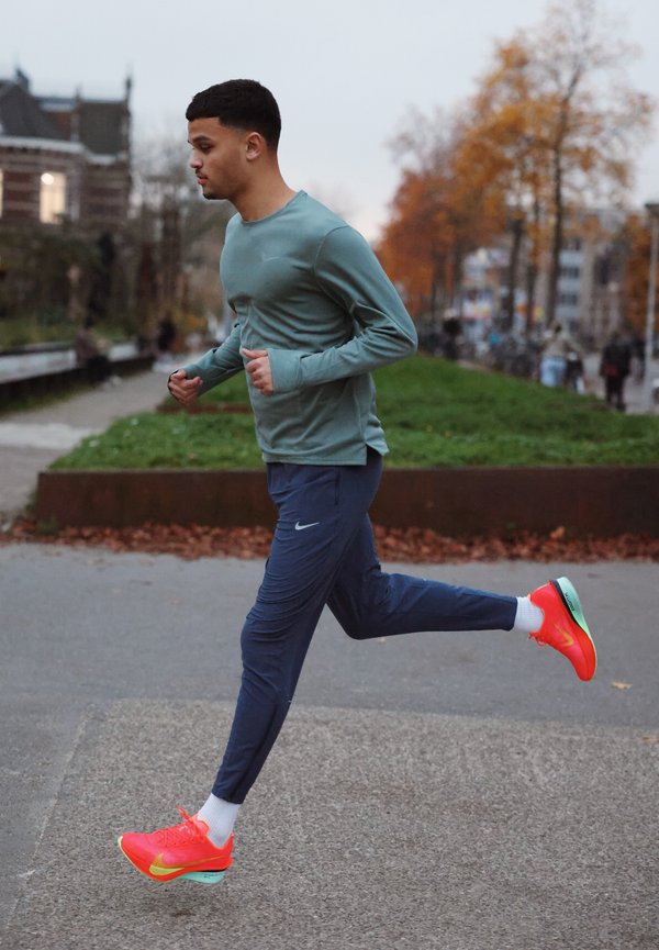 Sportos öltözék világoszöld hosszú ujjú pólóval és tengerészkék jogging nadrággal. Élénk narancssárga sportcipők türkiz részletekkel. Texturált út és zöld növényzet.