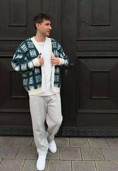 Cárdigan de punto con un patrón de cuadrícula en teal y blanco, con puños y escote en crema, combinado con una camiseta blanca y pantalones gris claro, de pie sobre la acera.