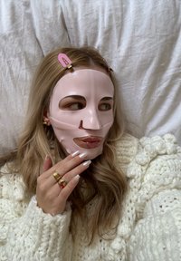 Femme aux longs cheveux portant un masque facial rose pâle, un pull blanc en maille épaisse, des bagues en or et des barrettes roses, allongée sur une couverture blanche.
