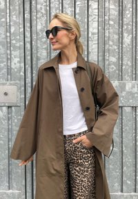 Brown oversized trench coat with large buttons, worn over a white t-shirt and leopard print trousers. Black sunglasses complete the outfit.