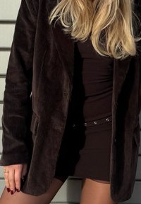 Rudas kordurovinis švarkas su pritaikyta forma, derinamas su aptempta juoda suknele, kurioje ant juosmens matomi metaliniai akcentai. Matomi lygūs paviršiai.