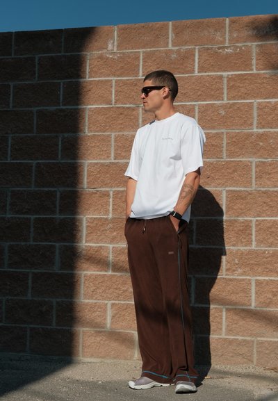 Camiseta blanca de manga corta, pantalones sueltos marrones con franjas laterales azules, acompañados de zapatillas grises. Texturas suaves, entorno urbano.