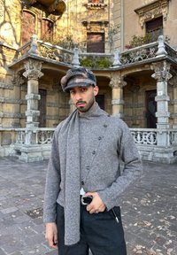 Homme portant un pull gris, une casquette plate à motifs et un pantalon noir, debout devant un bâtiment en pierre orné de colonnes et de balcons.