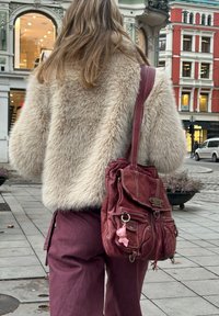 Jachetă pufoasă bej din blană artificială, pantaloni burgundy și o geantă de umăr din piele burgundy uzată, cu un charm decorativ. În aer liber, pe asfalt.