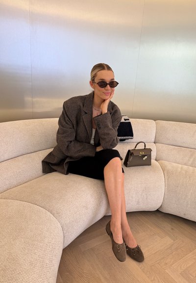 Blazer gris de gran tamaño, top negro ajustado, falda negra y zapatos de ante marrón. Pequeño bolso marrón con hardware dorado sobre un sofá texturizado beige.