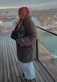 Manteau en fausse fourrure marron, manches longues, associé à un pantalon blanc à jambes larges. Tenant une pochette en cuir noir. Sol en bois en dessous.