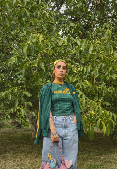 Chaqueta verde con cremallera y detalles amarillos sobre una camiseta a rayas verde de Adidas, jeans de tiro alto azul claro y un bolso de peluche colorido.