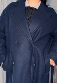 Persona che indossa un cappotto doppiopetto blu navy con le mani nelle tasche, sovrapposto a una giacca nera con bottoni e una camicia con colletto grigio.