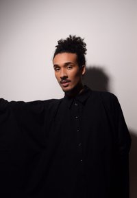 Chemise noire oversize en tissu lisse, avec un devant à boutons, un col pointu et des manches larges. Fond gris clair.