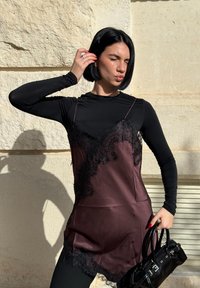 Žena s krátkymi čiernymi vlasmi pózujúca so zavret ými očami, oblečená v bordovej čipkovanej šatke cez čierne tričko s dlhým rukávom, držiaca čiernu kabelku.