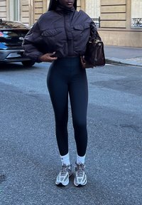 Brun cropped pufferjakke med to frontlommene, sammen med høytlivede svarte leggings. Grå og hvite joggesko, og en brun håndveske.