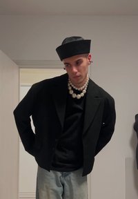 Zwarte oversized blazer met twee knopen, gedragen over een zwarte top. Grote parelketting en een zwarte, gestructureerde hoed. Lichtblauwe jeans.