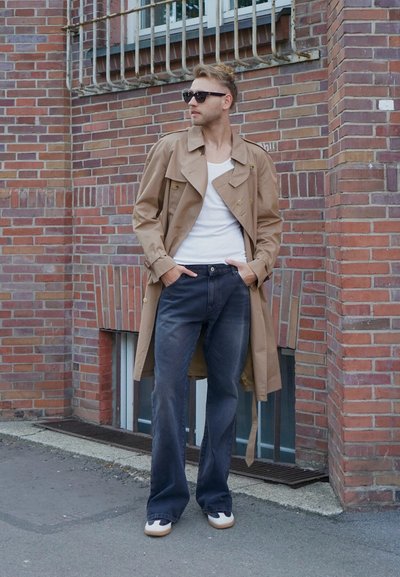 Gabardina beige con botones de doble botonadura, llevada sobre una camisa blanca y jeans oscuros. Gafas de sol negras y zapatos con suela blanca complementan el look.