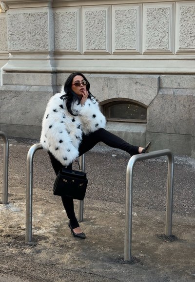 Mujer con abrigo de piel blanca con manchas y gafas de sol, posando con una pierna sobre un aparcabicicletas metálico frente a un edificio de piedra.