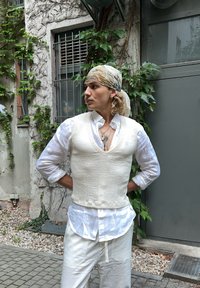Vesta albă texturată peste o cămașă albă cu nasturi, asociată cu pantaloni largi de culoare deschisă, pe un fundal de foliaj verde și un zid gri.