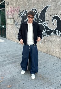 Sort cropped jakke over en hvid poloshirt, kombineret med løse blå denimjeans og hvide sneakers. Står foran en væg med graffiti.