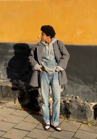 Un palton gri din lână peste un hanorac deschis la culoare cu un design grafic, blugi și pantofi în două nuanțe maronii pe un zid texturat galben și negru.