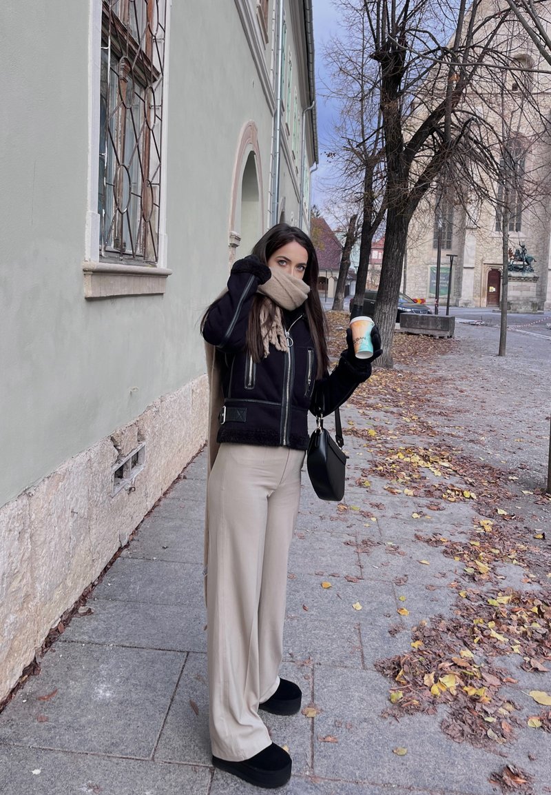 Μαύρο μπουφάν από shearling με φερμουάρ, μπεζ παντελόνι σε φαρδιά γραμμή, μαύρες πλατφόρμες, κρατώντας ένα φλιτζάνι καφέ, στέκεται σε έναν πλακόστρωτο δρόμο με πεσμένα φύλλα.